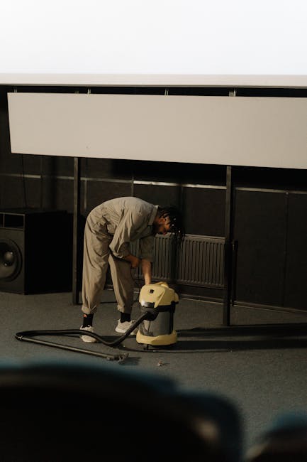 A man wearing a light grey jumpsuit is using a vacuum cleaner on a dark carpeted floor in an indoor setting, possibly a theater or auditorium with tiered seating visible in the background. The room fe
