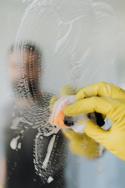 A person wearing yellow rubber gloves is cleaning a glass surface using a sponge and soapy water, creating a lather and streaks on the glass. The window or glass partition is partially obscured by the